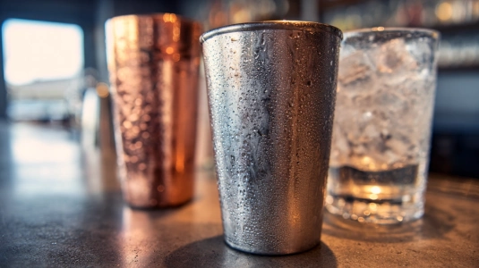 Rostfri stålshaker med kondensbildning bredvid en kopparshaker och ett tjockt mixglas