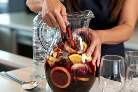 Glaskanna med sangria, skivad apelsin, citron och äpple samt en kanelstång, intill vinglas med is